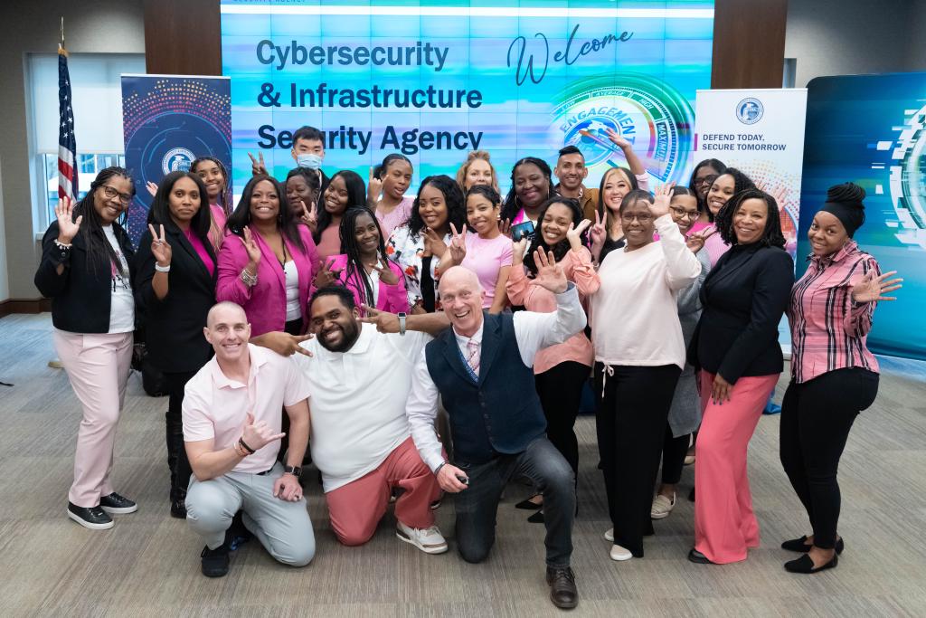 OCHCO staff photo (diverse group of men and women wearing pink) at Spring into CISA: In-Person Hiring Event. Group is in front of event branding and computer screens.